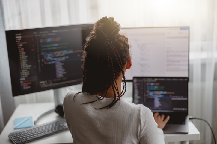 Rear of female web expert typing on computer looking at monitors sitting indoors, professional coder