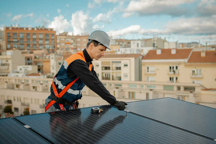 Worker in orange vest installing solar energy panels or photovoltaic cells on the rooftop of building in the city. Solar energy, Sustainable engineering, ecology and industrial concept. Horizontal